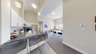 This single-floor, 5-bedroom, 2-bathroom unit offers a thoughtfully designed open-concept layout that maximizes functionality and shared living. The kitchen and living room flow seamlessly together beneath dramatically high ceilings, creating an airy, expansive atmosphere. The kitchen is well-appointed with stainless steel appliances — including a refrigerator, range, and dishwasher — white shaker cabinetry, a subway tile backsplash, dark countertops, and a breakfast bar with bar seating, ideal for casual dining or socializing. Adjacent to the kitchen, the combined dining and living area benefits from large windows that bring in generous natural light and views to the outdoors. The living room is generously proportioned with plush carpeting and ample space for multiple seating arrangements. Five private bedrooms provide individual retreats, each featuring carpet flooring and windows for natural light. The primary bedroom offers notable space and connects to hallway access toward the bathrooms. Both bathrooms are clean and functional, each featuring a full bathtub and shower combination. Two hallways provide organized circulation throughout the unit. The neutral color palette, soaring walls, and modern fixtures create a bright, contemporary atmosphere suited for shared or multi-occupant living.