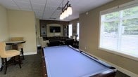 This single-floor recreational space is thoughtfully designed to serve as a social hub for leisure and entertainment. The centerpiece of the room is a full-sized billiards table with a striking light blue felt surface, illuminated by an elegant pendant light fixture featuring multiple frosted glass shades that hang from a drop-panel ceiling. The warm, neutral-toned walls create an inviting atmosphere throughout the space.

Adjacent to the billiards area is a comfortable lounge zone, furnished with wicker and upholstered seating arranged around a wall-mounted flat-screen television. Built-in dark wood cabinetry and shelving flank the TV wall, providing both storage and display space, adding a refined, finished look to the entertainment area. A desk or console surface is also integrated into this built-in unit, offering additional functionality.

Natural light flows generously into the room through multiple windows fitted with white horizontal blinds, offering views of surrounding greenery. The flooring features dark, patterned carpeting that adds texture and warmth underfoot. Overall, the space balances a casual, comfortable atmosphere with polished design details, making it well-suited for both relaxed lounging and active recreation.