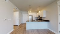This well-appointed single-floor residence showcases a thoughtfully designed open-concept layout with clean, contemporary finishes throughout. The kitchen serves as a central feature, boasting white cabinetry, dark quartz countertops, a large center island with an integrated sink and dishwasher, and a full suite of stainless steel appliances including a refrigerator, range, and microwave. Pendant lighting above the island adds a stylish touch, while recessed lighting ensures the space is well-lit throughout.

The living area is a standout, featuring expansive floor-to-ceiling corner windows and a set of glass-paneled French doors that flood the space with natural light and offer striking panoramic mountain views. Light wood-look flooring extends seamlessly through the main living and kitchen areas, enhancing the sense of openness.

The bedroom provides a comfortable retreat with plush carpeting, a window with blinds, and ample wall space. The bathroom is finished with a white vanity offering generous storage, a bold-framed mirror with bar lighting above, and a tub-shower combination, all set on wood-look tile flooring. The overall aesthetic is modern and cohesive, with a neutral color palette that creates a bright and inviting atmosphere throughout.