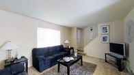 This three-floor residence offers a straightforward, functional layout designed for comfortable everyday living. The main level living room serves as a central gathering space, finished with neutral beige carpeting and clean, light-colored walls that create a bright and understated atmosphere. A large window fitted with a white roller blind allows natural light to fill the room, contributing to its open and airy feel. The space accommodates a full-sized sofa paired with a rectangular coffee table atop a textured area rug, providing a cozy and practical seating arrangement. A side table with a table lamp adds a warm accent to the corner of the room. A white-paneled interior door is visible along the back wall, offering access to an additional area on the main level. A white-railed staircase situated to the right of the living room provides access to the upper floors, with wall sconce lighting adding a decorative touch along the stairwell. Framed artwork displayed near the staircase adds a subtle decorative element to the space. Overall, the home presents a clean, neutral palette throughout, offering a versatile canvas well-suited to a variety of interior styles.