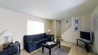 This three-floor residence offers a straightforward, functional layout designed for comfortable everyday living. The main level living room serves as a central gathering space, finished with neutral beige carpeting and clean, light-colored walls that create a bright and understated atmosphere. A large window fitted with a white roller blind allows natural light to fill the room, contributing to its open and airy feel. The space accommodates a full-sized sofa paired with a rectangular coffee table atop a textured area rug, providing a cozy and practical seating arrangement. A side table with a table lamp adds a warm accent to the corner of the room. A white-paneled interior door is visible along the back wall, offering access to an additional area on the main level. A white-railed staircase situated to the right of the living room provides access to the upper floors, with wall sconce lighting adding a decorative touch along the stairwell. Framed artwork displayed near the staircase adds a subtle decorative element to the space. Overall, the home presents a clean, neutral palette throughout, offering a versatile canvas well-suited to a variety of interior styles.
