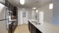 This single-floor residential unit offers a well-appointed, functional layout centered around a thoughtfully designed kitchen. The kitchen serves as a focal point of the space, showcasing rich dark espresso cabinetry that contrasts beautifully with the speckled stone countertops. A large kitchen island provides generous prep and workspace, fitted with a stainless steel double-basin sink and a sleek pendant light overhead, adding a contemporary touch to the room's ambiance. Stainless steel appliances, including a gas range, complement the modern aesthetic throughout. The flooring features warm wood-tone planks that run throughout the main living areas, lending the space a cohesive and inviting feel. Neutral light gray walls keep the interior bright and versatile, while flush and pendant lighting ensure the space remains well-lit throughout the day. A notable convenience feature is the in-unit laundry closet, discreetly tucked behind bifold doors and equipped with a side-by-side washer and dryer. Additional doors visible in the space suggest access to a main entry and utility or storage areas, reinforcing the practical and efficient design of this single-level home.