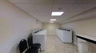 This shared laundry facility is a practical, well-organized space designed to accommodate multiple users simultaneously. The room features two long rows of top-load washing machines arranged along opposite walls, providing ample capacity for residents. The layout is efficient, maximizing the use of the available floor space while maintaining clear pathways between the machine rows.

The interior is finished with neutral beige walls and a drop ceiling equipped with recessed fluorescent panel lighting, ensuring bright, even illumination throughout the space. The light-toned vinyl tile flooring is easy to maintain and contributes to the clean, functional aesthetic of the room. A smoke detector is visible on the ceiling, indicating attention to safety standards.

A small seating area near the entrance features black folding chairs, offering a comfortable waiting area for users during wash cycles. The machines are neatly arranged in white cabinetry-style housings, presenting a uniform and tidy appearance. An informational notice is posted on the wall, likely providing usage guidelines or instructions. Overall, the space is clean, well-maintained, and purpose-built for convenient shared laundry use.