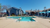 This residential community's outdoor amenity space is thoughtfully designed to offer residents a comprehensive range of leisure and recreational facilities. At the heart of the space is a generously sized swimming pool with crystal-clear blue water, complemented by an adjacent spa/hot tub, providing options for both active swimming and relaxation. The expansive sundeck surrounding the pool is outfitted with lounge chairs and bright blue market umbrellas, creating a welcoming atmosphere for outdoor recreation under open skies.

A covered pavilion with a wood-beam structure serves as a shaded gathering spot, adding architectural character to the outdoor environment. Nearby, an outdoor kitchen and grilling area is paired with dining tables and chairs, making the space well-suited for casual outdoor entertaining. Raised planter beds with ornamental landscaping and mature trees are integrated throughout, adding greenery and visual appeal to the hardscaped surroundings.

A children's playground is visible at the perimeter of the property, rounding out the family-friendly amenities on offer. The buildings surrounding the courtyard feature a distinctive blue-grey exterior with multi-level construction, pitched rooflines, and chimneys, creating an enclosed and cohesive community atmosphere. The entire space benefits from abundant natural light and a clear open sky, enhancing the overall outdoor experience.
