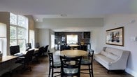 This single-floor space is designed with an open-concept layout that seamlessly integrates multiple functional zones. The kitchen area is highlighted by dark cabinetry with open shelving, a mosaic tile backsplash, stainless steel appliances, and a pendant chandelier that adds a warm focal point above a central dining table. The kitchen flows naturally into a secondary dining area in the foreground, where a round table is surrounded by dark wood chairs with patterned upholstered seats, creating an inviting spot for casual gatherings. To the left, a dedicated computer workstation area features multiple desks and ergonomic chairs, making the space well-suited for productivity alongside leisure. A comfortable sofa along the right wall provides a relaxed lounge option. Light-toned walls and hardwood flooring run throughout, creating a cohesive and airy atmosphere. Recessed lighting and natural light from windows enhance the sense of openness. The varied ceiling heights, including a raised soffit over the kitchen, add subtle architectural interest to the overall design.