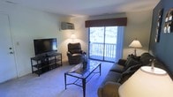 This single-floor space offers a welcoming and functional layout designed for comfortable everyday living. The living room serves as the heart of the space, featuring wall-to-wall carpeting that adds warmth underfoot. A large sliding glass door floods the room with natural light and provides direct access to a private balcony, framing pleasant views of the surrounding greenery. A valance-trimmed window treatment adds a classic decorative touch above the door. The living room is well-appointed with seating that includes a plush sofa and a leather recliner, centered around a sleek glass-top coffee table. A wall-mounted air conditioning unit ensures year-round climate comfort. Entertainment is catered for with a large flat-screen television set on a modern media console. Accent lamps positioned throughout the room contribute to a warm, inviting ambiance during evening hours. The overall design of the space balances practicality with comfort, making it a functional and pleasant environment. The connection between the interior living area and the outdoor balcony is a standout feature, allowing for natural ventilation and an easy indoor-outdoor lifestyle enhanced by the wooded backdrop visible through the glass door.