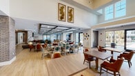 Located at 3171 US-280 E in Birmingham, Alabama, this single-floor amenity space offers a refined and welcoming environment designed with both comfort and style in mind. The open-concept layout flows seamlessly between distinct functional zones, united by warm wood-look flooring, intricate parquet tile inlays, and soaring double-height ceilings that lend an expansive, airy feel.

The dining area is a standout feature, accommodating multiple seating configurations with richly upholstered chairs in cognac leather and patterned fabrics. A dramatic coffered ceiling with geometric dark-framed panels adds architectural depth overhead, while curated wall art and metallic sculptural accents provide a polished finishing touch.

A full kitchen and bar area anchors one end of the space, outfitted with warm wood cabinetry, stainless steel appliances, a white countertop island, and tufted teal barstools beneath pendant lighting and a striking chandelier.

The lounge area features a sleek linear fireplace set into a marble surround, plush green velvet seating, and bold artwork — creating an inviting gathering space. A brick archway and exposed brick columns add textural contrast throughout.

Additional amenities include a billiards room with framed automotive artwork, a private meeting room with floor-to-ceiling windows overlooking outdoor grounds, and glass-enclosed flexible spaces for various activities.