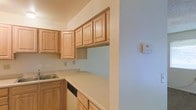 This single-floor space offers a clean and functional layout centered around a well-appointed kitchen. The kitchen features warm-toned raised-panel wood cabinetry that extends to the upper walls, providing generous storage capacity. Light-colored laminate countertops offer ample workspace and complement the cabinetry's natural finish. A built-in sink and a dishwasher are seamlessly integrated into the lower cabinet run, enhancing the kitchen's practicality. The walls are finished in a soft, neutral blue-gray tone that creates a calm and airy atmosphere throughout the space. A pass-through counter area connects the kitchen to an adjacent space, contributing to an open and accessible flow. A wall-mounted thermostat is conveniently located nearby, indicating a climate-control system for year-round comfort. The overall design is straightforward and efficient, prioritizing functionality with a cohesive, neutral color palette that makes the space feel bright and easy to personalize. The layout is well-suited for everyday living, with thoughtful storage solutions and durable finishes throughout.