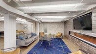This single-floor community amenity space at Hillsboro Village showcases a thoughtfully designed interior that blends industrial and contemporary styles throughout its generously sized layout. Exposed white-painted structural beams run across the ceilings, complemented by visible conduit and linear fluorescent lighting that reinforces the space's loft-inspired character. Whitewashed brick walls appear throughout, adding texture and warmth while unifying the various functional zones.

The open lounge area anchors one end of the space, featuring a gray sofa, tan leather accent chairs, a bold blue geometric area rug, and a wall-mounted flat-screen television positioned above a sleek fireplace set into a wood-paneled accent wall. Adjacent to this is a dedicated game room bearing the Hillsboro Village logo, equipped with a solid wood ping-pong table and natural light from low-set windows.

A dining zone with blue upholstered booth seating and white tabletops offers views of greenery through large windows. The expansive kitchen features white cabinetry, subway tile backsplash, stainless steel appliances, and a substantial stone-topped island with bar seating. Wood-look plank flooring flows throughout, tying each zone together seamlessly.