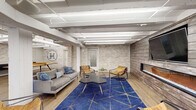 This single-floor community amenity space at Hillsboro Village showcases a thoughtfully designed interior that blends industrial and contemporary styles throughout its generously sized layout. Exposed white-painted structural beams run across the ceilings, complemented by visible conduit and linear fluorescent lighting that reinforces the space's loft-inspired character. Whitewashed brick walls appear throughout, adding texture and warmth while unifying the various functional zones.

The open lounge area anchors one end of the space, featuring a gray sofa, tan leather accent chairs, a bold blue geometric area rug, and a wall-mounted flat-screen television positioned above a sleek fireplace set into a wood-paneled accent wall. Adjacent to this is a dedicated game room bearing the Hillsboro Village logo, equipped with a solid wood ping-pong table and natural light from low-set windows.

A dining zone with blue upholstered booth seating and white tabletops offers views of greenery through large windows. The expansive kitchen features white cabinetry, subway tile backsplash, stainless steel appliances, and a substantial stone-topped island with bar seating. Wood-look plank flooring flows throughout, tying each zone together seamlessly.