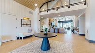 This single-story space makes an immediate impression with its dramatic vaulted ceiling clad in warm wood planking, accented by dark steel beams and a mezzanine-level glass panel that amplifies the sense of vertical height. The entry foyer sets an elegant tone with herringbone-patterned hardwood flooring, a bold circular accent table, patterned area rug, and crisp white shiplap walls adorned with framed artwork and gold-toned wall sconces.

The open-plan interior flows effortlessly from the welcoming lobby into a series of interconnected zones. A lounge area at the far end features floor-to-ceiling steel-framed glass walls that flood the space with natural light and provide views of an outdoor terrace furnished with seating. Curved lounge chairs and a round coffee table anchor this bright, airy retreat.

A large communal work table with a live-edge wood top and leather chairs serves as a central gathering point, positioned adjacent to glass-enclosed conference rooms visible in the background. A café-style kitchenette offers a commercial coffee station, white quartz countertops, slate-toned cabinetry, a blue mosaic tile backsplash, and a beverage refrigerator.

Lounge seating nooks with leather banquettes, bold abstract artwork, and bistro-style tables are tucked along interior walls, while a long corridor connects additional private spaces. The overall ambiance is modern farmhouse-meets-contemporary professional.