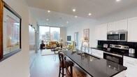 This stylish single-floor residence showcases a thoughtfully designed open-concept layout that seamlessly blends the kitchen, dining, and living areas into one cohesive, light-filled space. The kitchen is a standout feature, boasting crisp white cabinetry, a bold dark granite countertop island with bar seating, stainless steel appliances, and a modern high-arc faucet — combining both functionality and contemporary elegance. Recessed lighting throughout the ceiling provides consistent, warm illumination across the space.

The living area is anchored by floor-to-ceiling windows that flood the interior with abundant natural light and frame impressive views of the surrounding urban skyline. The open layout allows for a generous living and dining arrangement, complete with hardwood flooring that adds warmth and continuity throughout. A neutral color palette of whites and light grays forms the backdrop, complemented by warm furnishing tones that create an inviting atmosphere.

Overall, the space exudes a clean, modern aesthetic with a sophisticated urban sensibility, making it well-suited for those who appreciate contemporary design, open living, and a strong connection to the city environment.