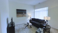 This two-story space presents a well-appointed interior designed with a clean, contemporary aesthetic. The living area is a standout feature, characterized by neutral white walls that create a bright, airy atmosphere. A large window dressed with sheer white curtains allows abundant natural light to fill the room, enhancing the sense of openness and space.

The living room is furnished with a dark upholstered sofa paired with a sleek glass-top coffee table supported by a sculptural base, reflecting a modern design sensibility. Soft, light-toned carpeting adds warmth underfoot and complements the room's overall neutral palette. Accent lighting is provided by table lamps positioned thoughtfully around the seating area. A large landscape-style canvas artwork depicting a coastal pier scene adorns one wall, serving as a focal point that adds depth and character to the space.

The overall ambiance is calm and understated, with a carefully curated mix of textures and tones that contribute to a polished, livable environment. The two-floor layout suggests additional living or private spaces beyond what is visible, offering functional versatility throughout the home.