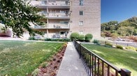 Located at 500 East Bruceton Road in Allegheny County, Pittsburgh, PA 15236, this single-level outdoor space offers a well-maintained and thoughtfully landscaped grounds area surrounding a multi-story brick residential building. The exterior features a concrete walkway lined with railing on both sides, providing safe and accessible passage along the length of the property. Colorful seasonal flower beds border the pathway, adding vibrant bursts of orange, yellow, and red to the lush green lawn. Mature deciduous trees provide generous canopy coverage throughout the grounds, offering natural shade and a sense of established character. A charming garden bench is positioned near a decorative flower arrangement, creating a peaceful outdoor retreat ideal for relaxation. A covered gazebo structure is visible toward the rear of the property, further enhancing the recreational outdoor amenities. The grounds overlook a parking area and an adjacent wooded hillside, framing the space with natural scenery. The residential building itself features multiple floors with private balconies, and ground-level access opens directly onto the landscaped grounds. The overall ambiance is calm, green, and inviting, reflecting a serene residential environment set within the rolling terrain characteristic of the greater Pittsburgh region.