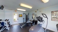 This single-story fitness room offers a functional and practical workout environment designed to accommodate a range of exercise needs. The space features light blue walls accented by white crown molding, creating a clean and open atmosphere. Overhead fluorescent lighting ensures the room is well-illuminated throughout the day.

The flooring transitions from rubber matting in the main workout area to tiled flooring near the entrance, providing both safety and durability underfoot. A glass-paned exterior door allows natural light to filter into the space while offering convenient direct access to the outdoors.

The room is equipped with a multi-function cable machine for strength training, an adjustable weight bench, a leg press or extension machine, and an Octane-brand elliptical trainer, catering to both resistance and cardiovascular workouts. Reference charts are mounted on the walls to assist users with exercise guidance.

A bulletin board near the entrance provides informational resources, and an emergency exit sign is clearly posted. Security fixtures are also present, reflecting attention to safety. Overall, the space delivers a straightforward and efficiently organized fitness environment.