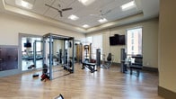 Located at 15 E Lane Ave in Columbus, Ohio, this well-appointed fitness center offers a comprehensive workout environment designed to accommodate a wide range of exercise needs. The single-floor facility features hardwood-style flooring throughout, a drop ceiling with recessed lighting, and ceiling fans that help maintain a comfortable atmosphere during workouts.

Large floor-to-ceiling windows flood the space with natural light while offering views of the surrounding Columbus cityscape, creating an open and energizing environment. The cardio section is equipped with multiple treadmills, elliptical machines, and a stationary bike, all positioned to take advantage of the natural light and outdoor views. A rowing machine provides additional low-impact cardio options.

The strength training area includes a multi-functional cable machine, a power rack, and a variety of selectorized resistance machines targeting different muscle groups. A dedicated free weights section features a full dumbbell rack backed by dark wood-paneled cabinetry, adding a refined aesthetic to the space. Wall-mounted flat-screen televisions are positioned throughout the facility for entertainment during workouts.

Full-length mirrors line portions of the walls, enhancing the sense of space and allowing for form monitoring. The facility also includes private restroom or changing room access. The overall design balances functionality with a polished, modern aesthetic suited for daily fitness use.