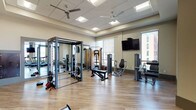 Located at 15 E Lane Ave in Columbus, Ohio, this well-appointed fitness center offers a comprehensive workout environment designed to accommodate a wide range of exercise needs. The single-floor facility features hardwood-style flooring throughout, a drop ceiling with recessed lighting, and ceiling fans that help maintain a comfortable atmosphere during workouts.

Large floor-to-ceiling windows flood the space with natural light while offering views of the surrounding Columbus cityscape, creating an open and energizing environment. The cardio section is equipped with multiple treadmills, elliptical machines, and a stationary bike, all positioned to take advantage of the natural light and outdoor views. A rowing machine provides additional low-impact cardio options.

The strength training area includes a multi-functional cable machine, a power rack, and a variety of selectorized resistance machines targeting different muscle groups. A dedicated free weights section features a full dumbbell rack backed by dark wood-paneled cabinetry, adding a refined aesthetic to the space. Wall-mounted flat-screen televisions are positioned throughout the facility for entertainment during workouts.

Full-length mirrors line portions of the walls, enhancing the sense of space and allowing for form monitoring. The facility also includes private restroom or changing room access. The overall design balances functionality with a polished, modern aesthetic suited for daily fitness use.