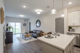 Located at 36 S New York Rd in Galloway, NJ, this single-floor unit offers a thoughtfully designed open-concept layout with a clean, contemporary aesthetic throughout. The kitchen serves as a central feature, equipped with crisp white shaker cabinetry, a white subway tile backsplash, stainless steel appliances including a dishwasher, microwave, and refrigerator, and a center island with a light countertop and pendant lighting above. The island provides seating and functions as a natural transition into the living area.

The living space is open and well-lit, with neutral-toned walls, recessed lighting, and durable wood-style flooring that extends throughout the unit. A sliding glass door in the living room fills the space with natural light and provides views of the surrounding landscape. The layout also includes a dedicated closet with wire shelving for ample storage.

The bathroom features marble-style porcelain tile flooring, a white vanity with undermount sink, a round mirror, chrome accents, and a tub-shower combination. A hallway connects the private spaces, maintaining a clear and functional flow. Overall, the unit presents a modern, well-appointed interior with consistent finishes and a bright, welcoming atmosphere.
