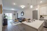 Located at 36 S New York Rd in Galloway, NJ, this single-floor unit offers a thoughtfully designed open-concept layout with a clean, contemporary aesthetic throughout. The kitchen serves as a central feature, equipped with crisp white shaker cabinetry, a white subway tile backsplash, stainless steel appliances including a dishwasher, microwave, and refrigerator, and a center island with a light countertop and pendant lighting above. The island provides seating and functions as a natural transition into the living area.

The living space is open and well-lit, with neutral-toned walls, recessed lighting, and durable wood-style flooring that extends throughout the unit. A sliding glass door in the living room fills the space with natural light and provides views of the surrounding landscape. The layout also includes a dedicated closet with wire shelving for ample storage.

The bathroom features marble-style porcelain tile flooring, a white vanity with undermount sink, a round mirror, chrome accents, and a tub-shower combination. A hallway connects the private spaces, maintaining a clear and functional flow. Overall, the unit presents a modern, well-appointed interior with consistent finishes and a bright, welcoming atmosphere.