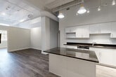 Located at 24 Mt Vernon St in Lynn, MA, this 1-bedroom, 1-bathroom unit showcases a striking industrial-loft aesthetic with soaring ceilings, exposed structural beams, and white-painted ductwork that add architectural character throughout the open floor plan. Wide-plank hardwood-style flooring runs seamlessly across the main living areas, anchoring the space with a warm, contemporary feel.

The kitchen is a standout feature, boasting a large center island topped with dark granite countertops, crisp white cabinetry, a classic subway tile backsplash, and a full suite of stainless steel appliances including a range, microwave, dishwasher, and refrigerator. Stylish pendant lighting hangs above the island, complemented by a track lighting system overhead. The open layout connects the kitchen directly to the living area, creating a versatile and social living environment.

The living area offers generous open space, a built-in closet for storage, and access to a staircase with an elegant railing detail. The bathroom is finished with large-format dark slate-style floor tiles, a white vanity with an undermount sink, and clean, neutral walls that provide a calm, functional space. Overall, the unit delivers a modern urban living experience with thoughtful design details throughout.