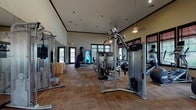 This fitness center offers a comprehensive workout environment housed within a single-level space designed to accommodate a wide variety of exercise needs. The room features notably high ceilings with crown molding details, creating an open and airy atmosphere that prevents the space from feeling cramped despite its substantial equipment offerings. Recessed lighting and pendant fixtures provide consistent illumination throughout, complemented by abundant natural light streaming through multiple large, dark-framed windows lining the perimeter walls.

The flooring consists of durable, patterned carpet tiles in warm tones, providing cushioning underfoot across the entire workout area. Equipment is well-distributed throughout the space and includes a robust selection of Precor strength machines, cable systems, a functional trainer, treadmills, ellipticals, stationary bikes, and a dedicated free weight station with a tiered dumbbell rack and weight bench. Full-length mirrors along one wall enhance the sense of space and allow users to monitor their form during exercise.

A wall-mounted television provides entertainment, while a beverage vending machine offers added convenience. Direct door access to an outdoor area is available, further enhancing the overall functionality and accessibility of this well-appointed fitness facility.
