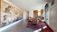 This single-floor lobby on N Fenwick Ave in Portland, OR, is a well-designed communal entry space that balances modern architecture with warm, inviting details. The layout divides naturally into distinct functional zones while maintaining a cohesive aesthetic throughout.

The main lounge area makes an immediate impression with a large-scale, wall-spanning historical mural rendered in sepia tones, depicting Portland's early cityscape and maritime heritage. This anchors the room as both an art installation and a nod to the neighborhood's history. Recessed ceiling lighting illuminates the space evenly, complementing the natural light that pours in through expansive floor-to-ceiling windows along the street-facing wall, offering open views of the surrounding neighborhood.

The seating area features tall-backed leather wingback chairs arranged on a bold red and charcoal patterned area rug, creating a comfortable and visually striking gathering space. Adjacent to this is a built-in library nook with warm wood shelving stocked with books, flanked by a cushioned L-shaped banquette and a flat-screen television, offering a more intimate retreat within the broader lobby.

Durable large-format tile flooring extends through the entry corridor, where a full wall of black residential mailboxes provides a practical and organized service area. The wood-paneled ceiling accents near the entry add warmth, tying the design together cohesively.