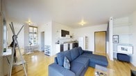 This single-floor space offers a well-designed open-plan layout that seamlessly connects the living and kitchen areas into one cohesive, functional environment. The living area is anchored by a generous L-shaped sectional sofa in a cool navy blue, complemented by warm honey-toned hardwood flooring that runs throughout the space, adding a sense of warmth and continuity. Crisp white walls and smooth white ceilings reflect natural light streaming in from a large window, keeping the interior feeling airy and bright. The kitchen is neatly integrated into the space, featuring dark cabinetry contrasted against white appliances, including a full-size refrigerator, stove, and microwave. Open shelving provides practical storage and display opportunities, while the clean layout ensures ease of movement. A compact corner workspace near the window offers a dedicated area for desk use, bathed in natural light. The main entrance is framed by a solid wood door, adding a touch of warmth to the entryway. Overall, the space delivers a modern, efficient, and inviting atmosphere suitable for everyday living.