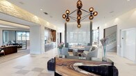 This upscale amenity lounge is a masterclass in contemporary design, seamlessly blending natural materials with modern elegance. The grand entrance features black-framed floor-to-ceiling glass doors and walls, flooding the space with natural light and offering clear sightlines to the surrounding exterior. Light-toned tile flooring in a subtle checkered pattern anchors the open layout throughout.

At the heart of the lounge, a dramatic double-sided linear fireplace set against a dark stone surround serves as a striking focal point, with a large wall-mounted television above. Plush, oversized sectional seating invites relaxation, complemented by live-edge wood coffee tables that add organic warmth. A sculptural bronze globe chandelier and artistic pendant lighting throughout enhance the refined atmosphere.

A dedicated coffee and refreshment bar, framed by textured natural stone accent walls and rich dark wood cabinetry, provides a functional yet stylish amenity. Adjacent, a dining area with a long communal wood table beneath an array of perforated metallic pendant lights overlooks expansive windows with open views. A separate lounge nook, dressed in warm tones and contemporary artwork, rounds out the space. A glass-railed staircase provides access to additional levels, completing this thoughtfully curated environment.