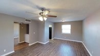 This single-floor space offers an open layout with a practical and functional design throughout. The main living area is finished with rich, dark hardwood-style flooring that extends across the space, complementing the neutral gray walls and crisp white baseboards. A ceiling fan with a built-in light fixture is centrally mounted, providing both airflow and ambient lighting, while a secondary flush-mount light fixture adds additional illumination near the window. A single window along the far wall allows natural light to filter into the room, contributing to a bright and welcoming atmosphere.

The open floor plan connects the living area directly to the kitchen, which is visible through a pass-through or open doorway. A black refrigerator can be seen in the kitchen area, hinting at included appliances. Notably, in-unit laundry is accessible from the main living space, with a visible washer positioned near the kitchen entry — a practical convenience. The textured ceiling adds a subtle design detail throughout. The overall layout prioritizes functionality with a clean, move-in-ready aesthetic suited for everyday living.