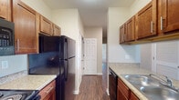 This 2-bedroom, 2-bathroom residence at The Concord Point in Memphis, Tennessee presents a well-organized single-story floor plan designed for comfortable everyday living. The galley-style kitchen is outfitted with rich medium-toned wood cabinetry, granite-look countertops, a stainless steel sink, and black appliances including a refrigerator, electric range, and built-in microwave, all set atop durable wood-style flooring that extends throughout the main living areas. The open transition between the kitchen and living area creates a connected feel, with neutral-toned walls providing a versatile backdrop. Both bedrooms feature plush carpeting, ceiling fans with light fixtures, and windows with blinds that allow for natural light. One bedroom includes a built-in desk and shelving, offering a dedicated workspace. Each bathroom is appointed with rich wood-tone vanities, granite-look countertops, framed mirrors with lighted vanity bars, and wood-style flooring. Accessibility features such as grab bars are present in one of the bathrooms. The overall aesthetic is clean and consistent, with warm finishes and a neutral palette that create a welcoming, functional living environment.