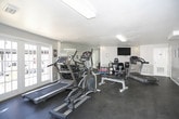 This single-floor fitness center offers a practical and functional space designed to support a variety of workout routines. The gym is equipped with multiple cardio machines, including treadmills and elliptical trainers, as well as a weight bench and a dumbbell rack, providing options for both cardio and strength training. A large wall-mounted mirror on one side of the room expands the sense of space and allows users to monitor their form during exercise. A flat-screen television is mounted on the wall, offering entertainment during workouts, while rolled towels and a small storage shelf are conveniently available nearby. The flooring throughout is durable rubber gym matting in dark charcoal, well-suited for high-traffic fitness use. Natural light flows generously into the space through a set of large French doors, which also provide direct views of the exterior. A Coca-Cola vending machine is positioned near the entrance for easy refreshment access. Motivational signage adorns the walls, contributing to an energetic atmosphere. The walls feature a two-tone paint scheme — white on the upper half and gray on the lower — giving the room a clean, modern appearance. Fluorescent and recessed lighting ensure the space remains well-lit throughout. Multiple doorways connect to adjacent utility or restroom areas, adding to the overall convenience of the facility.