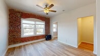 Located in the Koreatown neighborhood of Los Angeles, this single-floor unit at 200 S Kenmore Ave offers a blend of classic character and practical modern updates. The main living area immediately draws attention with its striking exposed brick accent wall, complete with an arched window surround that frames views of the surrounding streetscape and palm trees beyond. Light wood-style flooring runs throughout, complementing the bright, white walls and creating a warm, airy atmosphere. A ceiling fan overhead provides both comfort and style. Natural light flows generously through multiple windows, enhancing the open feel of the space. A separate bedroom features its own ceiling fan, built-in closet shelving, and ample room to furnish comfortably. The kitchen is equipped with slate-blue cabinetry, granite-style countertops, a stainless steel sink with a modern faucet, and black appliances including a refrigerator and range. The bathroom is clean and efficiently laid out, featuring a white vanity with framed mirror, toilet, and a tiled shower area. The overall ambiance of the unit is bright and inviting, with character-rich architectural details balanced by updated finishes throughout.