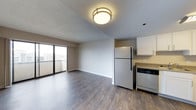 This single-floor unit offers an open-concept layout that seamlessly connects the living area and kitchen into one cohesive space. The flooring throughout is finished in a warm, medium-toned hardwood-style material that adds a sense of continuity and polish to the interior. A flush-mount ceiling light with a modern metal ring frame provides overhead illumination, complementing the natural light that enters through a large sliding glass door. This glass door leads directly to a private balcony, offering outdoor access and elevated views of the surrounding area. The living space features clean, neutral-toned walls that provide a versatile backdrop for a variety of décor styles. The kitchen is positioned at one end of the open floor plan and includes white upper cabinetry, a full-size refrigerator, and a dishwasher, offering practical functionality within a compact footprint. A countertop workspace adds to the kitchen's utility. The textured ceiling and simple, uncluttered design of the space contribute to a bright and functional ambiance that suits everyday living.