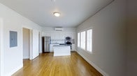 This single-floor space showcases an open-concept layout that seamlessly integrates the kitchen and living areas. The kitchen is outfitted with white shaker-style cabinetry, a decorative tile backsplash, and a suite of stainless steel appliances, including a refrigerator, gas range, and built-in microwave. A central island provides additional counter space and serves as a natural dividing point between the kitchen and the adjoining living area. The warm-toned hardwood floors extend throughout the space, adding a sense of continuity and warmth to the overall design. The walls are finished in a clean, bright white, complementing the light and airy atmosphere. A wall-mounted mini-split unit ensures efficient climate control. Natural light enters generously through a set of windows fitted with white blinds along one wall, brightening the interior without compromising privacy. The layout is open and functional, offering a versatile space well-suited for both everyday living and entertaining. A doorway visible on the left side of the space suggests additional rooms or storage beyond the main open area.