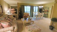 This single-floor space showcases a warm and inviting cottage-inspired design that seamlessly blends the living and dining areas into one cohesive, open layout. The living area is furnished with floral-upholstered sofas and an ottoman, centered around a whitewashed wooden coffee table adorned with fresh floral arrangements, lending a classic, homey atmosphere. Soft, neutral carpeting runs throughout, complementing the warm sage-green walls and earthy tones of the décor. Tall topiary plants and decorative greenery add a natural, lively touch to the corners of the room. Large windows dressed in sage-green drape panels allow abundant natural light to filter in, offering pleasant views of the surrounding wooded landscape and a glimpse of an outdoor deck or balcony. The dining area flows naturally from the living space and features a white rectangular dining table paired with matching white ladder-back chairs, accommodating a comfortable number of guests. Overhead, an elegant chandelier with candelabra-style lighting adds a refined focal point to the dining space. A white display cabinet and decorative side tables complete the room, reinforcing the cohesive, cottage-chic aesthetic throughout. Overall, the space exudes a relaxed yet tastefully appointed ambiance ideal for both everyday living and casual entertaining.
