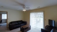 This single-floor residential space offers a welcoming and functional layout designed for comfortable everyday living. The living room serves as the central gathering area, furnished with a dark leather sofa and a wooden coffee table with lower storage shelf, all set atop plush carpeted flooring that adds warmth to the space. A ceiling fan with integrated light fixture is mounted overhead, providing both air circulation and ambient lighting. One of the standout features of the living area is the full-height sliding glass door dressed with vertical blinds, which allows natural light to filter into the room and provides a view of the outdoor surroundings. The neutral, light-toned walls create a clean and versatile backdrop throughout the space. A wall-mounted HVAC vent is visible, indicating a climate control system integrated into the structure. The overall ambiance is practical and cozy, with a straightforward design that prioritizes livability and ease of use. The single-floor layout ensures convenient accessibility throughout the entire space.