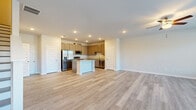 This two-story space is designed with a clean, contemporary aesthetic, featuring light hardwood-style flooring that runs consistently throughout both floors, contributing to a cohesive and airy feel. The main level centers around an open-concept layout where the kitchen flows seamlessly into the living area. The kitchen is well-appointed with taupe shaker-style cabinetry, granite countertops, a central island with additional seating and storage, and a full suite of stainless steel appliances including a refrigerator with ice and water dispenser, range, microwave, and dishwasher. Recessed lighting illuminates the space efficiently.

The upper level offers multiple rooms, including bedrooms with neutral-toned walls, ceiling fans, and natural light from windows. Closet space is well-considered, with one room featuring a walk-in closet outfitted with wire shelving systems for organized storage, and another with a standard reach-in closet. A dedicated laundry room on the upper level includes washer and dryer hookups and wire shelving above. A smart thermostat is also present. Throughout, the design prioritizes neutral tones, practical storage, and functional layouts.