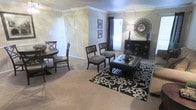 This single-story space showcases an open-plan layout that seamlessly connects the dining area and living room, creating a cohesive and functional environment. Crown molding runs along the ceiling throughout, adding a refined architectural detail to the otherwise neutral palette of soft gray and beige walls. The flooring is wall-to-wall carpet in a warm beige tone, providing comfort underfoot across both areas. The dining area, positioned to one side, features a round wooden table accompanied by classic X-back chairs, suited for everyday dining. A set of vertical blinds covers what appears to be a sliding door, offering access to an outdoor area while allowing flexible light control. The living area centers around a tufted leather ottoman serving as a coffee table, flanked by a pair of upholstered accent chairs. A bold black-and-white patterned area rug anchors this seating zone. Natural light filters in through a curtained window, brightening the space. A dark wood media console along one wall provides storage and display space, decorated with a decorative sunburst mirror and lamp, adding visual interest to the room.