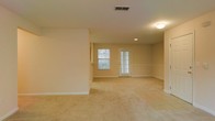 This single-floor space features an open living area finished in a clean, neutral palette. The room is dressed in wall-to-wall beige carpeting that extends throughout, complementing the warm, light-toned walls and white baseboards. Recessed lighting and a flush-mount ceiling fixture provide consistent illumination across the space, while a window and an adjoining glass-paneled exterior door along the far wall invite natural light into the interior. The door features decorative glass panes, adding a subtle architectural detail to an otherwise understated design. The open layout creates a generous sense of space, offering flexibility in how the area can be arranged and used. A partial wall division subtly defines separate zones within the room without interrupting the overall open feel. The ceiling incorporates a visible HVAC vent, indicating a central air system. The overall ambiance is calm and neutral, providing a blank canvas that accommodates a wide range of interior design preferences. The combination of soft carpeting, natural light, and a straightforward layout makes this a functional and adaptable living space.