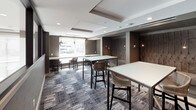 This contemporary lounge space is thoughtfully designed to accommodate both focused work and casual social interaction. The single-floor layout is unified by a patterned geometric carpet in muted grey and blue tones, complementing the space's sophisticated, neutral color palette of warm greys and dark taupe walls.

Central to the space are multiple white high-top tables paired with woven rattan-style bar chairs, providing flexible seating arrangements throughout. A built-in counter along the windowed wall offers an additional workspace, illuminated by a series of decorative pendant lights in varied metallic forms that add visual interest overhead. Large windows at this workstation flood the area with natural light and frame views of a pool terrace and surrounding urban skyline.

A standout feature is the recessed booth area, which showcases floor-to-ceiling tufted upholstered panels in a warm taupe fabric, flanked by adjustable wall-mounted reading lights. This alcove creates an intimate setting within the larger open environment. Tray ceiling details with recessed lighting add architectural depth, while a glass partition wall connects the space visually to an adjacent room, enhancing the sense of openness and scale.