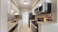 This single-floor residence offers a well-organized layout across a comfortable footprint, as illustrated by the 3D floor plan. The galley-style kitchen is a functional highlight, featuring white shaker cabinetry, granite countertops, stainless steel appliances including an electric range, over-the-range microwave, dishwasher, and refrigerator, along with a decorative mosaic tile backsplash and light ceramic tile flooring. The kitchen connects efficiently to the main living and dining areas.

The living area is bright and open, showcasing light gray wood-style flooring and neutral warm-toned walls. Large sliding glass doors flood the space with natural light and provide direct access to an outdoor balcony with views of surrounding greenery. An additional room, likely a dining area or secondary living space, mirrors this aesthetic with another set of sliding doors.

The bedrooms are carpeted for comfort, finished with the same neutral tones and fitted with standard six-panel doors. The bathroom features a full-sized soaking tub with a tiled shower surround, chrome fixtures, a grab bar, and wood-style flooring, offering a clean, functional design throughout.