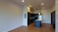 This single-floor space showcases a contemporary open-concept design that seamlessly blends the living and kitchen areas into one cohesive, functional environment. The kitchen is a standout feature, equipped with dark espresso cabinetry that contrasts beautifully against light stone countertops. A central island with a navy blue base provides additional workspace and casual seating, while stainless steel appliances — including a refrigerator, range, and microwave — lend a sleek, modern finish. A tiled backsplash adds a subtle decorative touch behind the range. Recessed lighting is installed throughout the ceiling, complemented by elegant pendant lights that hang above the kitchen island, creating focused task lighting with a stylish flair. The floors are finished in warm-toned hardwood, adding a sense of warmth and continuity across the open space. The living area is generously proportioned with clean white walls and tall ceilings, contributing to an airy, uncluttered ambiance. A utility panel is neatly recessed into the wall, maintaining the clean aesthetic. The overall design balances practicality with modern style, making this a well-appointed and inviting space.
