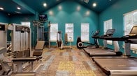 Located at 9500 Brightway Ct in Henrico, Virginia, this well-equipped fitness center offers a comprehensive workout environment designed to support a wide range of exercise routines. The space is immediately distinguished by its bold teal walls and vaulted ceiling, creating an energizing atmosphere accentuated by recessed lighting throughout.

The main workout area is thoughtfully organized to accommodate both cardio and strength training. A row of cardio machines — including treadmills, an elliptical, and a stair climber — lines one side of the room, positioned near windows that allow natural light to filter in. On the opposite side, a cable and weight machine station with adjustable benches provides a dedicated space for resistance training. A full-length mirror along one wall enhances the sense of openness and allows for form monitoring during workouts.

A well-stocked free weight rack offers a range of dumbbells, while a separate functional fitness area features stability balls, resistance bands, yoga mats, and a body composition scale — all neatly organized near louvered storage closets. The mosaic-patterned carpet tiles in earth tones and muted yellows add a dynamic visual element underfoot.

Multiple windows provide views of the surrounding greenery, contributing to a bright and motivating environment. A wall-mounted television adds entertainment options for those using cardio equipment, making this fitness center a fully functional and inviting space for daily exercise.