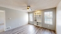 This single-floor unit offers a clean, functional layout designed for comfortable everyday living. The main living area is bright and open, featuring durable wood-look vinyl plank flooring, soft neutral gray walls, and a ceiling fan overhead. A standout built-in shelving unit with varied compartments serves as both a decorative focal point and practical storage solution, while multiple windows fitted with blinds invite an abundance of natural light throughout.

The kitchen is well-appointed with crisp white cabinetry, light countertops, and black appliances including a range and refrigerator. A peninsula-style breakfast bar provides a convenient pass-through to the living area, enhancing the open flow between spaces. The layout is efficient, offering ample counter and cabinet space for meal preparation.

A separate carpeted living or flex room features a sliding glass door that opens to a private outdoor patio area, offering a seamless indoor-outdoor connection. The textured ceiling and ceiling fans are consistent throughout the main areas.

The bathroom is neatly finished with a wide vanity, light countertops, white cabinetry, a large mirror with overhead lighting, and standard fixtures — functional and clean in presentation. Overall, the space presents a cohesive, neutral palette well-suited for a variety of furnishing styles.