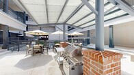 This outdoor grill and lounge area in Lafayette, Louisiana, is designed as a versatile communal gathering space set within a multi-story building's open-air courtyard. The space is divided into two distinct zones that complement each other seamlessly.

The grill station is positioned beneath a structured pergola roof supported by bold gray columns, providing shade over a stainless steel gas grill mounted atop a decorative red brick base. Prep tables flank the grill, and umbrella-shaded dining tables and chairs are arranged nearby, offering a comfortable setting for outdoor meals. Railings and views toward an adjacent pool area further enhance the ambiance of this section.

The adjacent open courtyard lounge features a bright, airy atmosphere bathed in natural light. Woven outdoor chairs with green cushions are arranged in a conversational grouping around a central coffee table, creating an inviting social space. Concrete bench-style seating blocks are also present throughout. The surrounding architecture showcases neutral-toned horizontal siding, dark-framed doors, and structural columns that continue upward to wrap-around upper-level balconies with metal railings.

Together, the two zones create a well-rounded outdoor amenity space that balances functional grilling and dining with relaxed lounge seating, all framed by clean, contemporary architectural details.