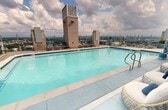 Located in Nashville, this 720-square-foot rooftop pool and amenity space offers a resort-style experience high above the city. The centerpiece is a large, sparkling aquamarine pool surrounded by a spacious tiled sun deck outfitted with modern loungers in teal and gray, perfect for relaxing while taking in panoramic views of the Nashville skyline. An adjacent covered lounge area provides a comfortable indoor-outdoor retreat, featuring a striking stacked stone fireplace column with a mounted flat-screen television above. The lounge is furnished with contemporary outdoor sofas and accent chairs accented with blue and teal throw pillows, creating a cohesive, stylish aesthetic. Architectural details throughout include clean-lined gray and white panel walls, polished concrete-style flooring, and floor-to-ceiling glass doors that connect the interior lounge to the open-air deck. Lush green plantings in built-in and freestanding planters add natural contrast to the modern palette. The covered lounge area is equipped with recessed lighting and ceiling-mounted fixtures, ensuring usability at any hour. Wicker accent chairs and additional seating zones provide versatile gathering options, making this rooftop destination ideal for both relaxation and socializing against a breathtaking Nashville backdrop.