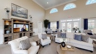 This clubhouse is designed to provide a refined and comfortable community gathering space, highlighted by an expansive great room with dramatically tall ceilings adorned with crown molding. Arched transom windows near the roofline flood the interior with natural light, complemented by large French doors and floor-to-ceiling windows framed by rich navy blue drapes that offer views of an outdoor pool area. The great room is thoughtfully furnished with plush neutral sofas, accent chairs, and layered area rugs, creating multiple intimate seating arrangements suited for socializing or relaxing. Wainscoting lines the lower walls throughout, adding a classic architectural detail that ties the space together. A built-in media console with shelving anchors one side of the room, while lush indoor trees and potted plants add a fresh, vibrant touch. A separate dining area accommodates a full-sized dark wood table with upholstered seating and a wall-mounted television, ideal for group gatherings or meetings. Cozy nook seating, highlighted by a pair of navy blue accent chairs, warm floor lamps, and decorative artwork, offer quieter retreat spaces within the clubhouse. The overall ambiance is elegant yet approachable, blending traditional design elements with comfortable, contemporary furnishings.