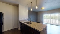 This single-floor residential unit offers a thoughtfully designed layout that balances functionality and modern style. The kitchen serves as a central highlight, anchored by a large island with granite countertops and a deep double-basin sink illuminated by a trio of pendant lights hanging at varying heights. Dark espresso cabinetry — including upper glass-front cabinets — pairs with a full suite of black appliances, including a refrigerator, electric range, and microwave. Rich dark hardwood-style flooring runs throughout the kitchen and living areas, lending warmth and visual continuity. The living area is open and airy, featuring a sliding glass door with vertical blinds that invites natural light and provides access to an outdoor balcony. An accent wall in a deep charcoal tone adds a modern design touch to the space. The bedroom is carpeted for comfort and features a large window with outdoor greenery views and a walk-in closet area with built-in shelving. The bathroom is clean and well-equipped, featuring a tub-shower combination, a granite-topped vanity with dark wood cabinetry, and a large mirror. Neutral tones throughout the unit create a cohesive and inviting atmosphere.