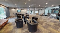 This two-story clubhouse in Ann Arbor, Michigan offers a well-rounded combination of social, recreational, and functional spaces across both levels. The main level is anchored by a spacious lounge area with patterned carpet flooring, recessed lighting, and structural columns that define distinct seating zones. Clusters of leather club chairs surround glass-topped coffee tables, creating comfortable gathering spots throughout the open floor plan. A standout brick fireplace wall serves as a dramatic focal point, adding warmth and character to the lounge. Wall-mounted televisions and high-top bar seating provide additional options for casual use. A contemporary kitchen area features stainless steel appliances, dark cabinetry, and an island with bar-height seating beneath a teal-accented ceiling detail. The fitness center spans a dedicated area and includes cardio equipment, free weights, and a weight bench, with a separate room for strength training. A staircase connects to the upper level, where a bright office and meeting space features light hardwood flooring, large windows with views of surrounding greenery, and glass partitions that allow natural light to flow freely. An octagonal skylight element enhances the upper landing, adding architectural interest and illuminating the transition between floors.