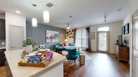 This single-floor residence offers a thoughtfully designed open-concept layout that seamlessly connects the living, dining, and kitchen areas. The living room is bright and welcoming, featuring warm hardwood-style flooring, large windows with stylish patterned drapes that invite abundant natural light, and a ceiling fan overhead. A decorative arrangement of gold circular wall accents adds a distinctive artistic touch to the space.

The kitchen is sleek and modern, showcasing rich dark cabinetry paired with light quartz countertops, a subtle tile backsplash, and a full suite of stainless steel appliances including a refrigerator, dishwasher, and range. A breakfast bar with pendant lighting overhead provides casual seating and serves as a natural divider between the kitchen and living areas.

The bedroom is a serene retreat, finished with plush carpet flooring, soft neutral tones, and generous natural light filtering through sheer curtains. A ceiling fan and ample storage space contribute to the room's comfort and functionality.

The bathroom features a wood-framed vanity mirror, dual-sink countertop, hardwood-style flooring, and a full tub-shower combination. An in-unit laundry area is conveniently accessible nearby. Together, these spaces deliver a cohesive, contemporary living environment with modern finishes throughout.